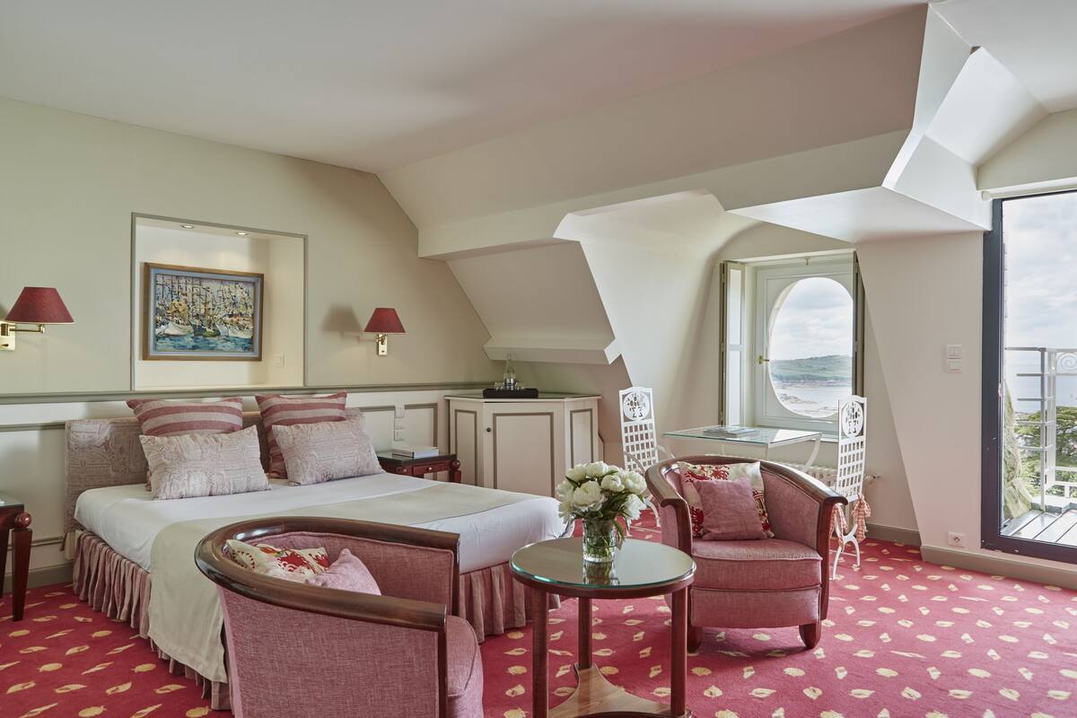 A hotel room with a large bed, a sitting area with a table and chairs, a window with a view, and a painting on the wall.