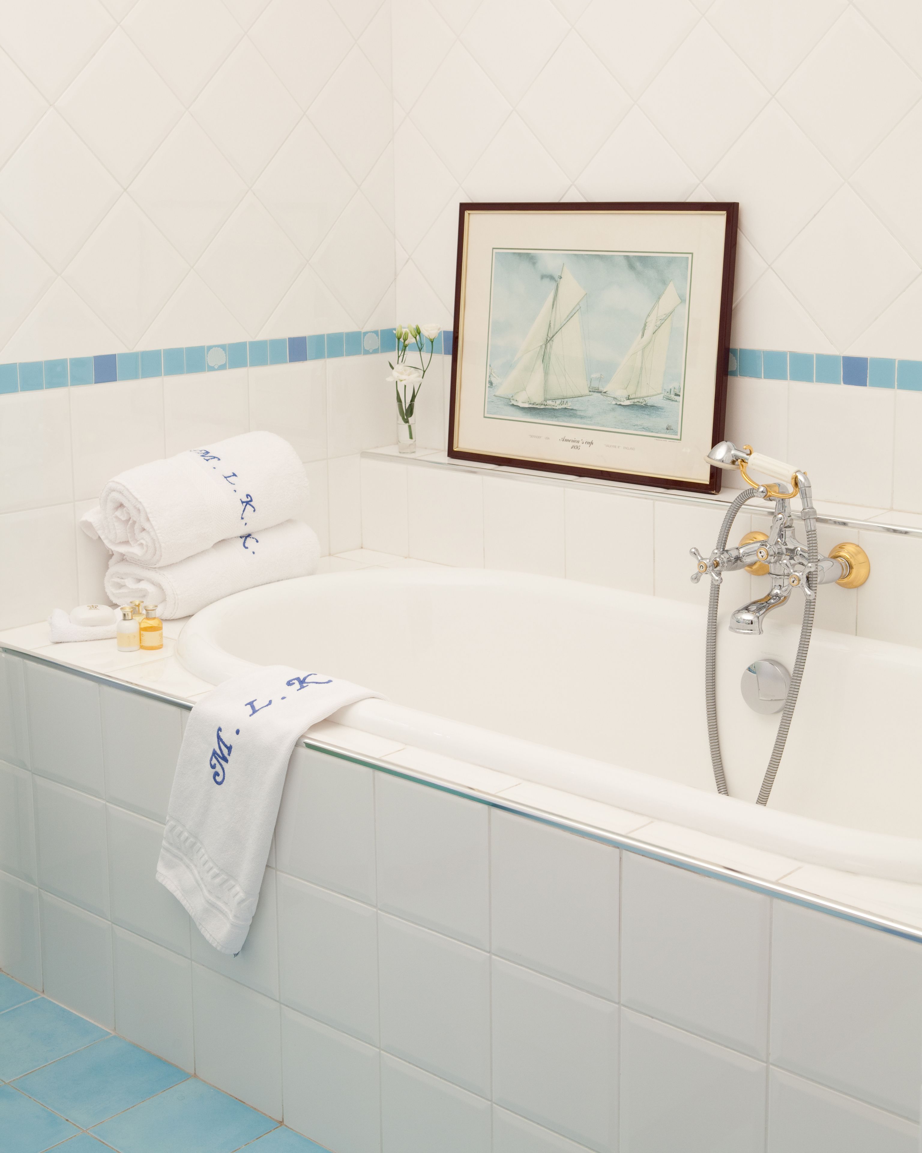A bathroom with a bathtub, a framed painting of sailboats, and a small vase with flowers.