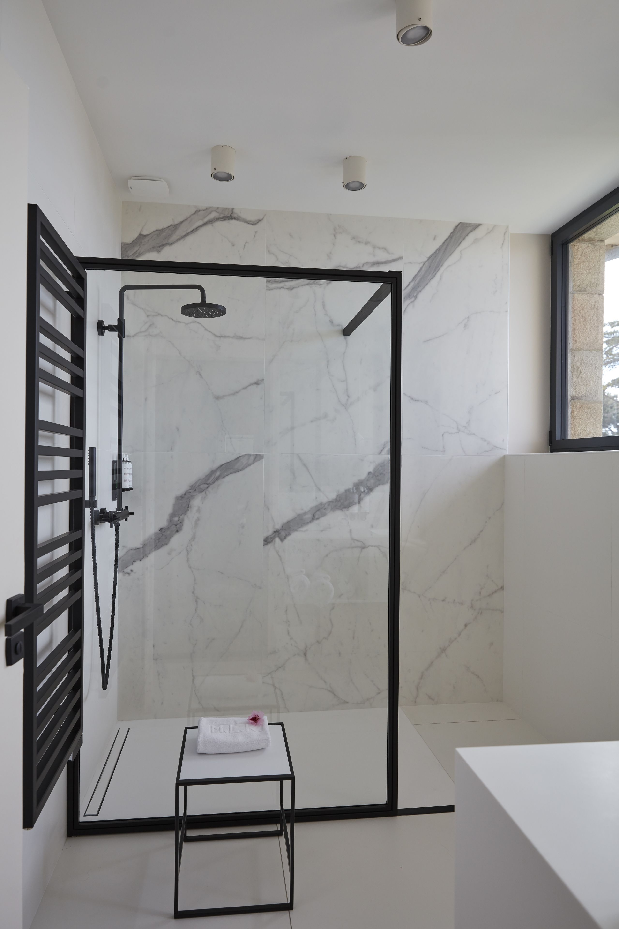 Un baño moderno con pared de ducha de mármol, cerramiento de vidrio con marco negro, lavamanos blanco y una pequeña mesita con una toalla.