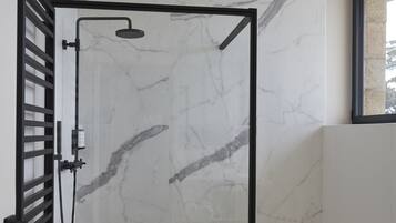Un baño moderno con pared de ducha de mármol, cerramiento de vidrio con marco negro, lavamanos blanco y una pequeña mesita con una toalla.