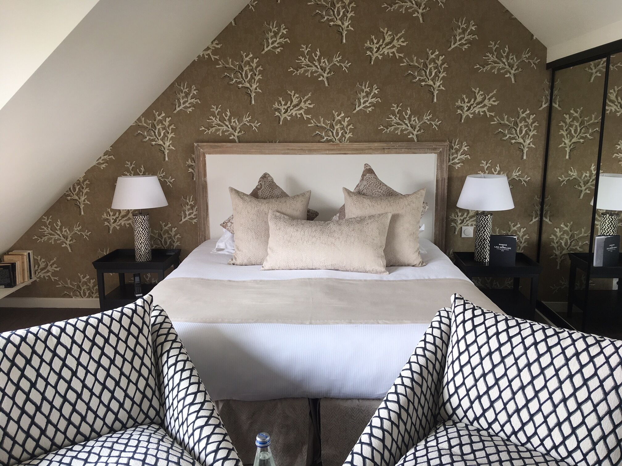 A bedroom with a sloped ceiling, a large bed with multiple pillows, two bedside tables with lamps, and a patterned wall.