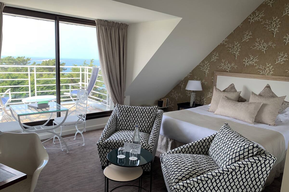 A hotel room with a bed, two patterned armchairs, a small table, and a balcony with a view of the sea.
