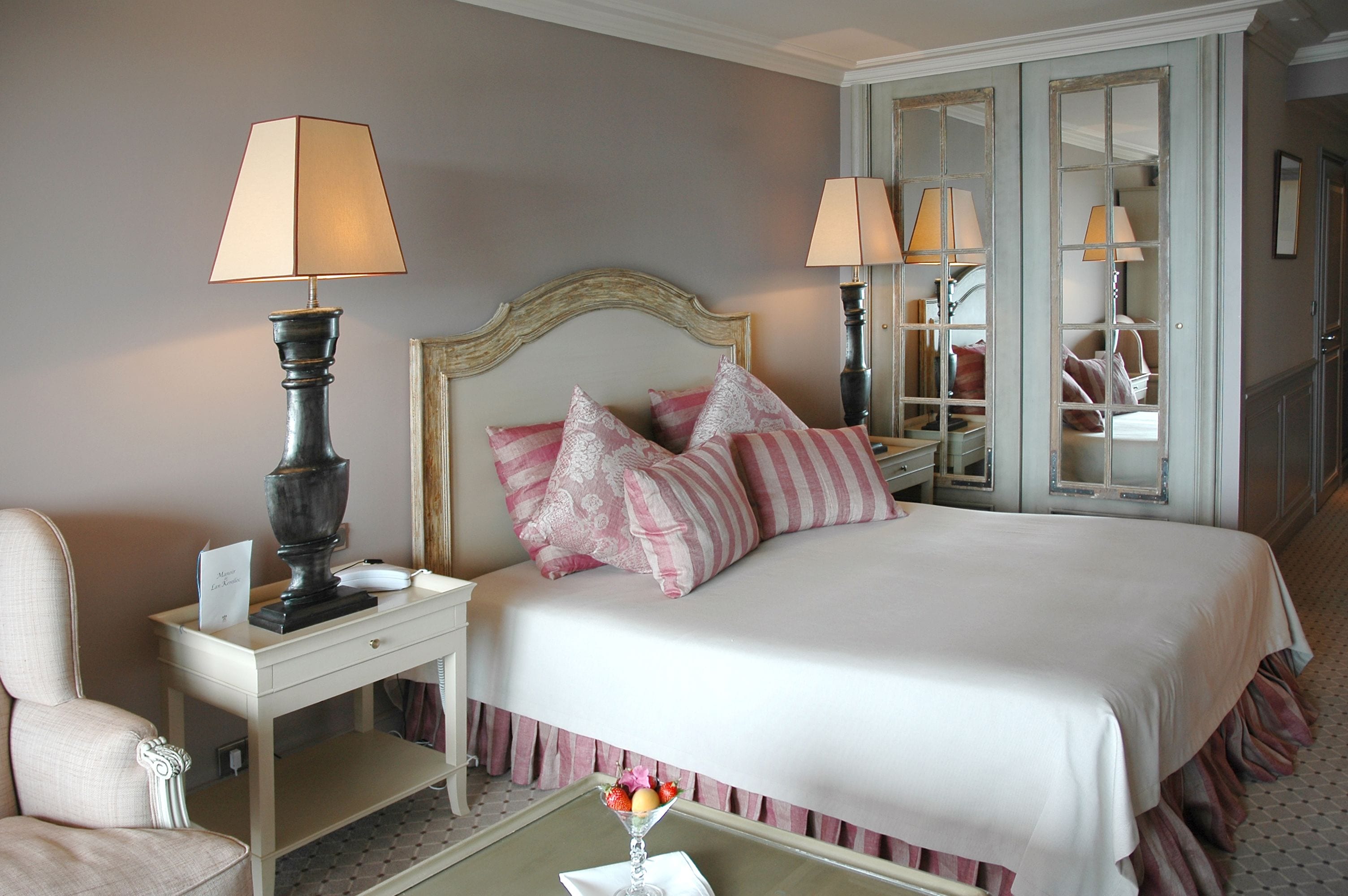 A hotel room with a large bed, two bedside lamps, a nightstand, a chair, and a small table with a fruit bowl.