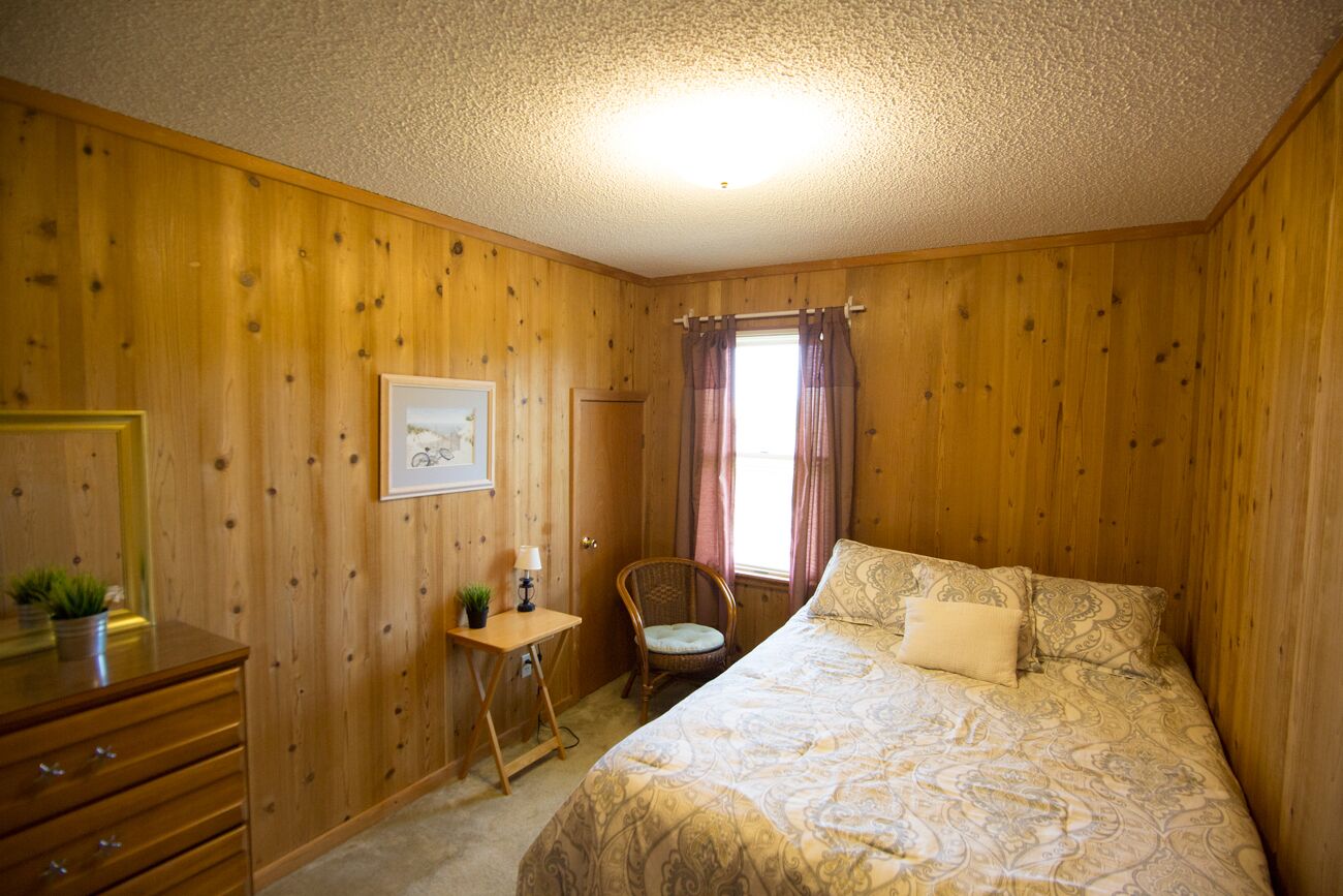 A wooden-paneled room with a bed, a desk, a chair, and a window with curtains.