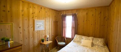 A wooden-paneled room with a bed, a desk, a chair, and a window with curtains.