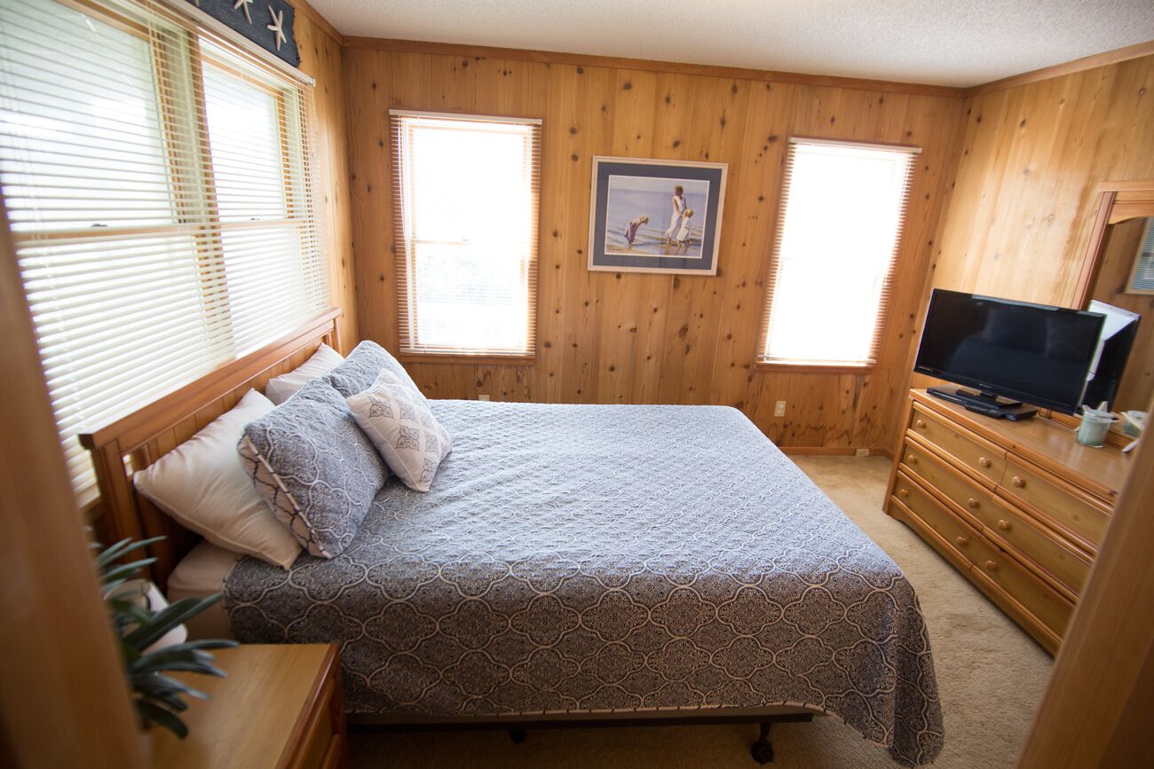 Une chambre à coucher avec un lit, des murs en bois, une commode, un téléviseur et deux fenêtres.