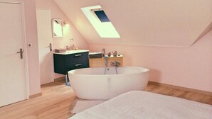 A bedroom with a large bathtub, a skylight, a vanity with a mirror, and a door. - La Ferme d'Antan (Mons-Boubert)