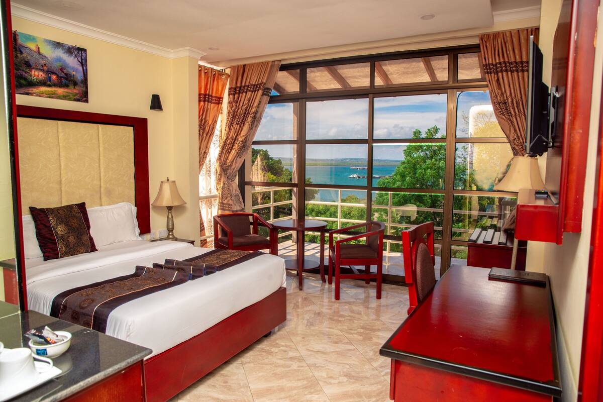 A hotel room with a large bed, a desk, a chair, and a view of the sea.