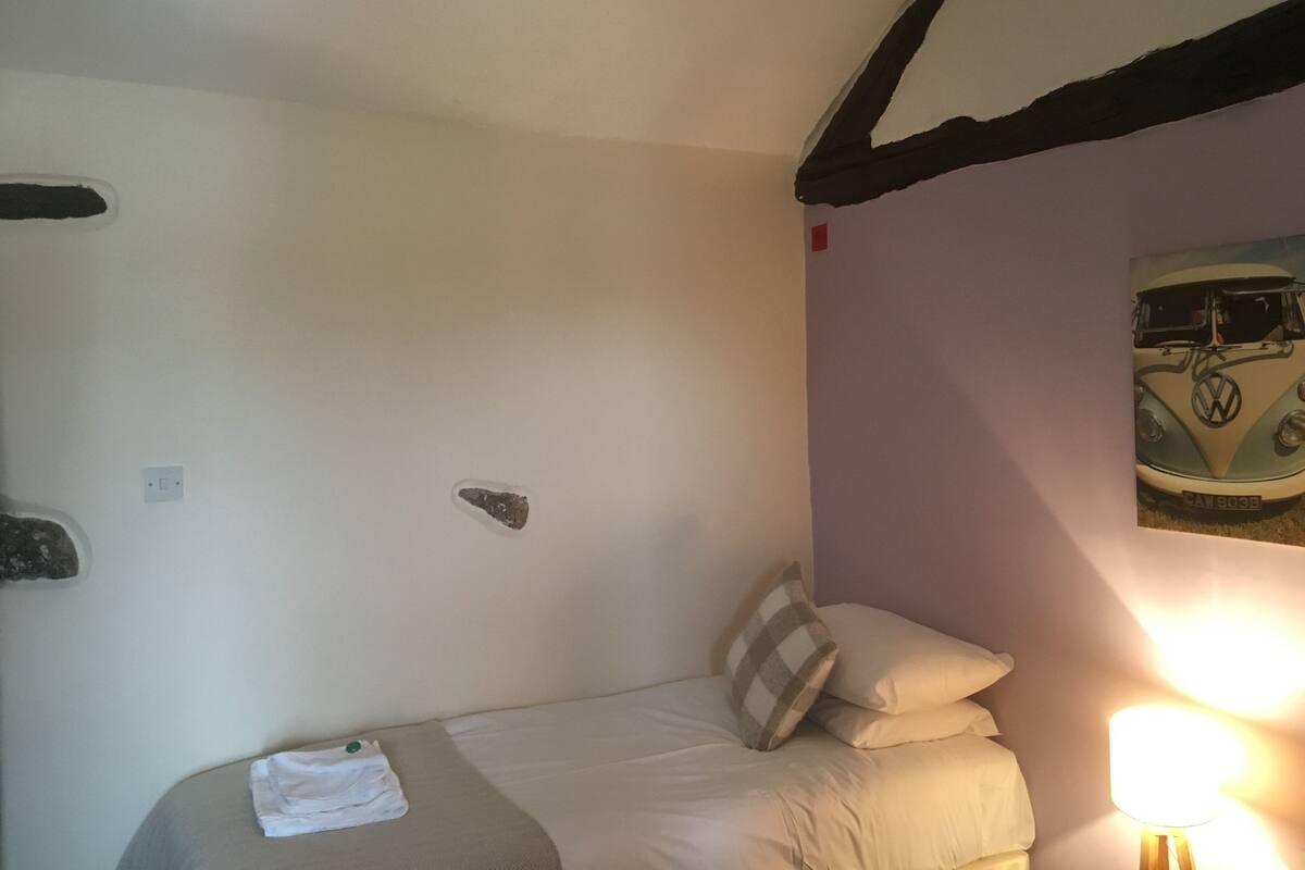 A bedroom with a bed, pillows, a lamp, a painting of a vintage car, and a small decorative item on the wall.