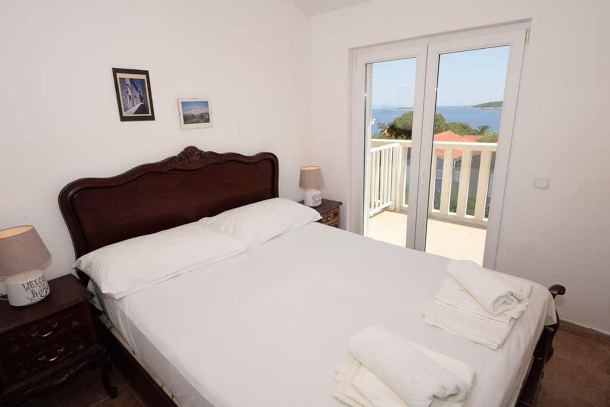 Ein Schlafzimmer mit Bett, Nachttisch, Lampe und Meerblick durch eine Balkontür.