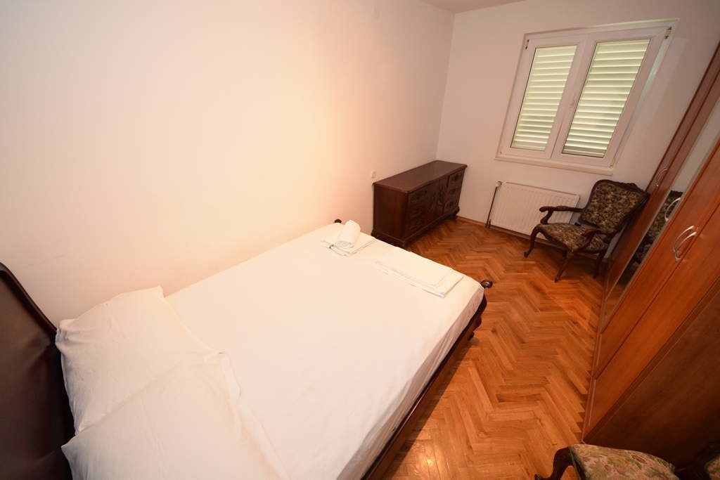 Ein Schlafzimmer mit Holzboden, einem Bett mit weißen Bettwäsche, einer Holzkommode, einem Stuhl und einem Fenster mit Jalousien.