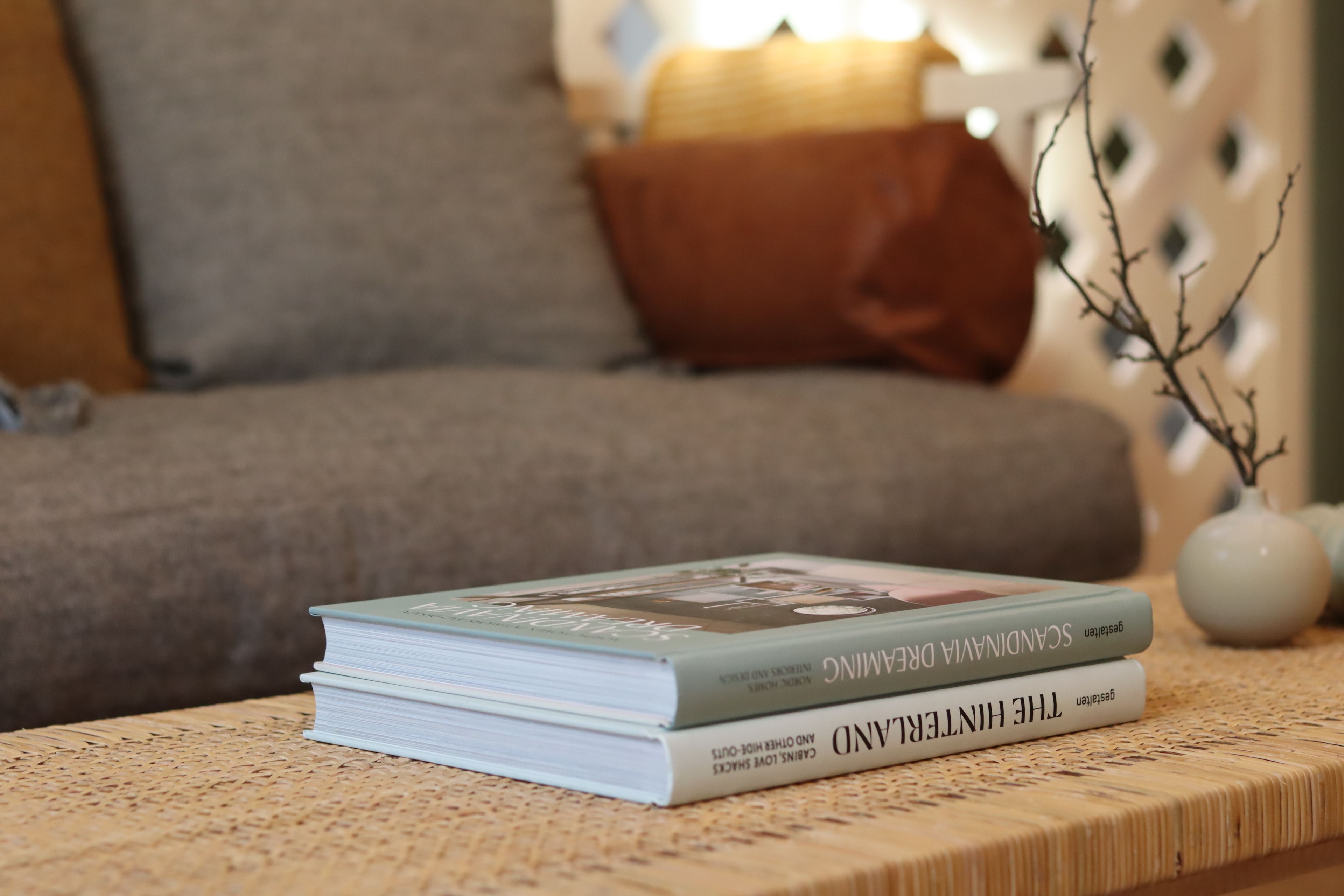 A stack of books on a woven table with a sofa and decorative vase in the background.