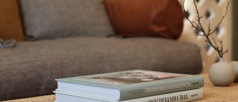A stack of books on a woven table with a sofa and decorative vase in the background.