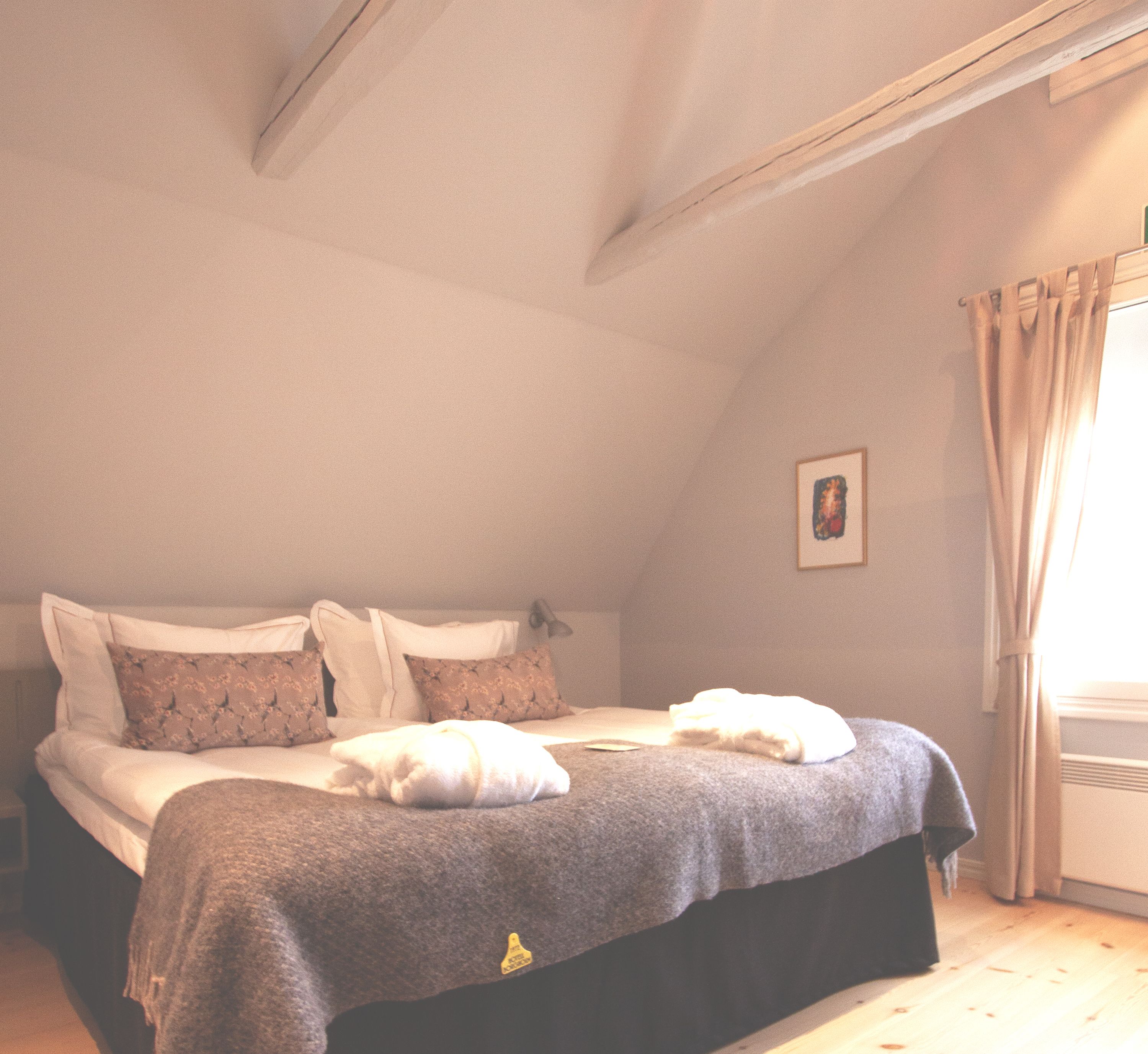 Ein Schlafzimmer mit Bett, Kissen, Decke, einem Bild an der Wand und einem Fenster mit Vorhängen.