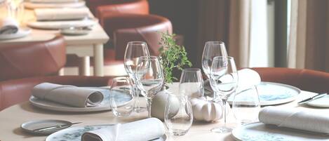A dining area with round tables set for two, each with folded napkins, clear wine glasses, and plates.