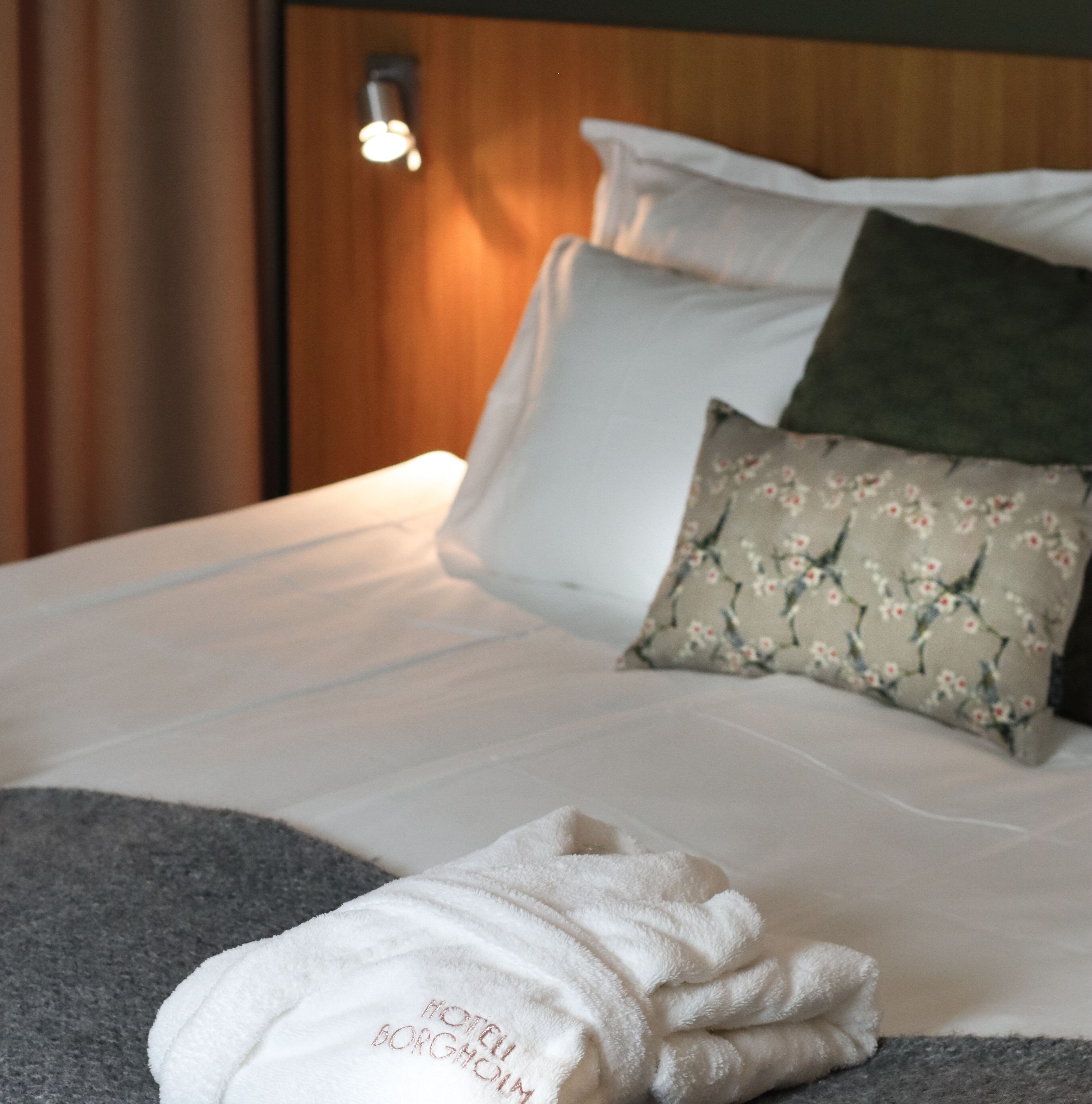 Un lit bien fait, avec des draps blancs, un coussin à motifs floraux et une serviette pliée sur laquelle est brodé le nom « HÔTEL BORGÅM ».
