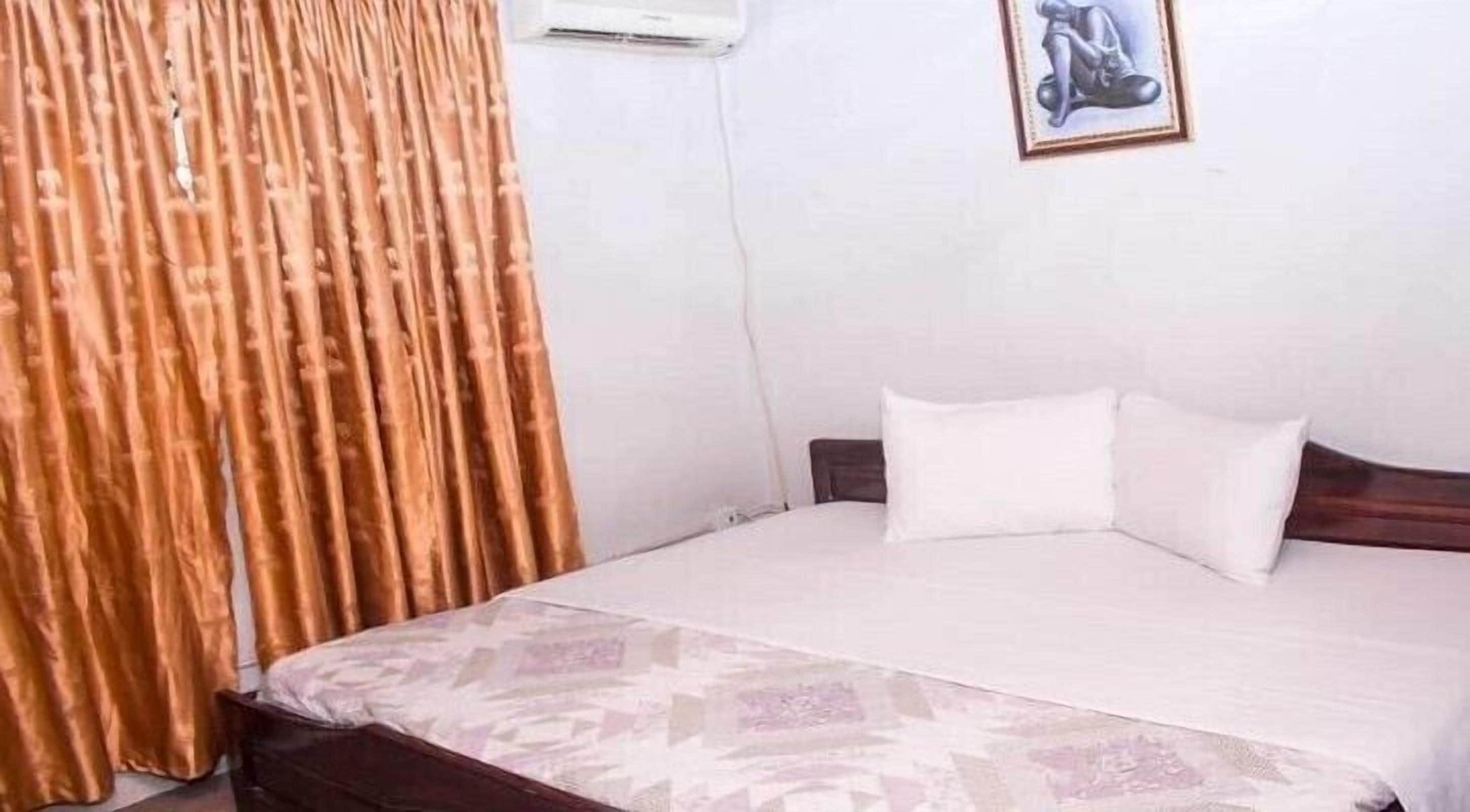 A bedroom with a bed, a patterned bedspread, a wooden headboard, a white pillow, and a framed picture on the wall.