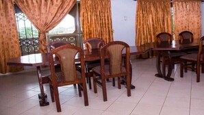A dining area with wooden tables and chairs, patterned curtains, and a view through a window with a metal grille. - Vinny Hotel (Sekondi-Takoradi)