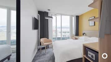 Hotellihuone, jossa on sänky, työpöytä, tuoli, televisio ja suuri ikkuna, josta avautuu näkymä kaupunkiin.