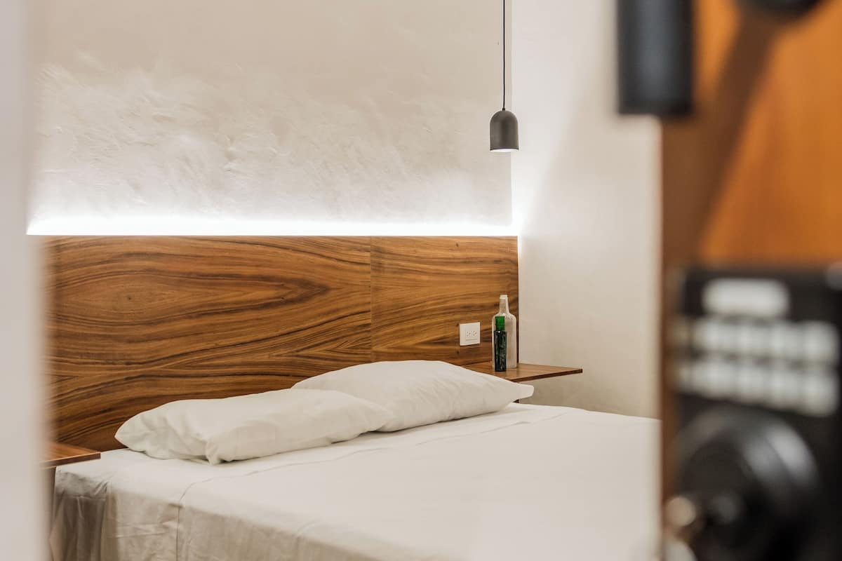 A bedroom with a wooden headboard, a bed with white bedding, a bedside table with a bottle, and a hanging light fixture.