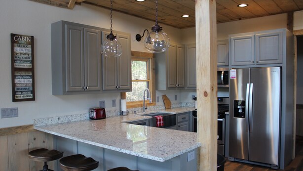 A kitchen with grey cabinets, a stainless steel refrigerator, a granite countertop, and hanging pendant lights.