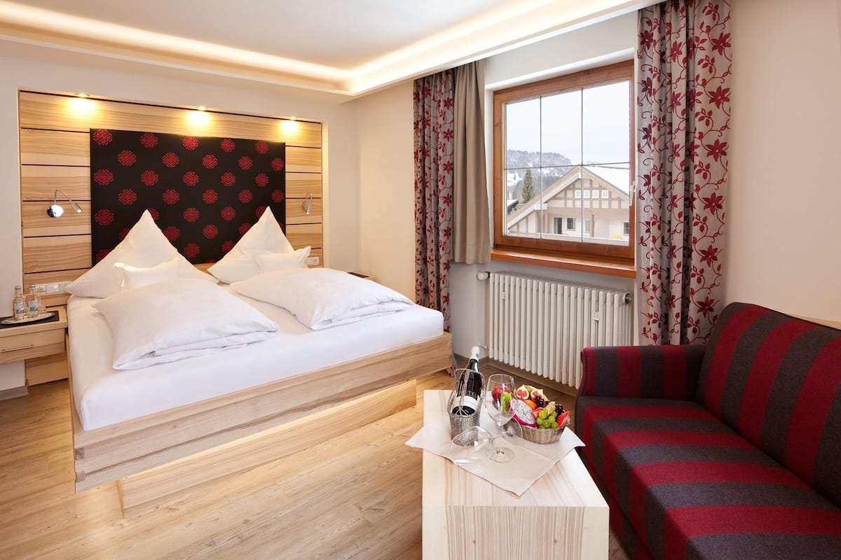 Une chambre d’hôtel avec un lit, une tête de lit en bois, une table de chevet, un canapé rouge et gris, une fenêtre avec des rideaux et une vue sur les bâtiments à l’extérieur.