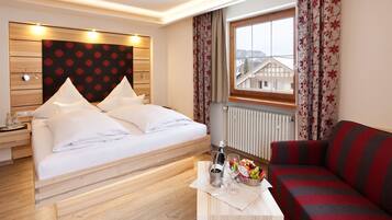 A hotel room with a bed, a wooden headboard, a nightstand, a red and grey sofa, a window with curtains, and a view of buildings outside.