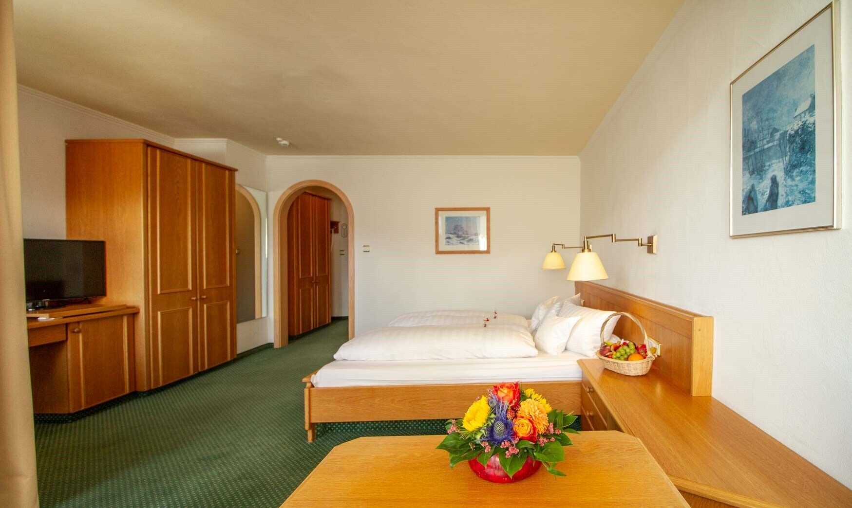 A hotel room with a bed, a desk with a television, wooden wardrobe, and a small table with a fruit basket.