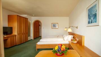 A hotel room with a bed, a desk with a television, wooden wardrobe, and a small table with a fruit basket.