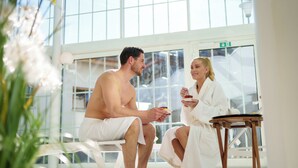 A man and a woman in a spa setting with a large window, a small wooden stool, and a plant. - Hotel Filser (Oberstdorf)