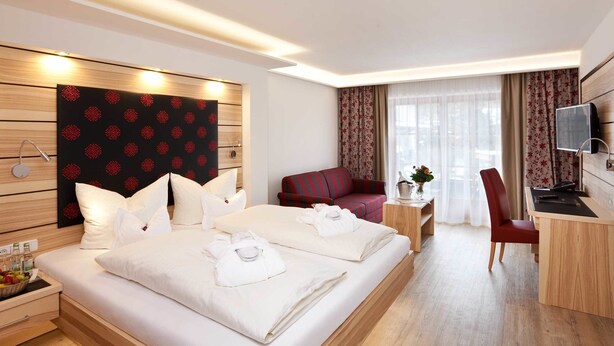 A hotel room with a large bed, a desk, a chair, a TV, and a window with curtains. - Hotel Filser (Oberstdorf)