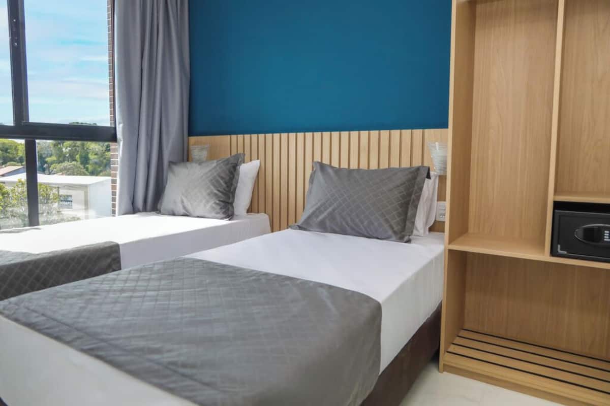 Ein Hotelzimmer mit zwei Betten, einem Holzkopfende, einer eingebauten Ablage und einem Fenster mit Blick auf Gebäude.