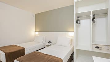 A hotel room with two single beds, a bedside table with a phone and lamp, a wardrobe with hanging clothes, and a desk with a chair.