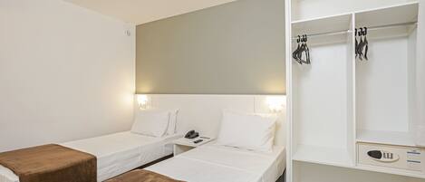 Una habitación de hotel con dos camas individuales, una mesita de noche con teléfono y lámpara, un armario con ropa colgada y un escritorio con silla.