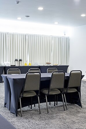 Un salón de conferencias con una mesa larga, seis sillas y un montaje tipo buffet con platos, vasos y salsas.