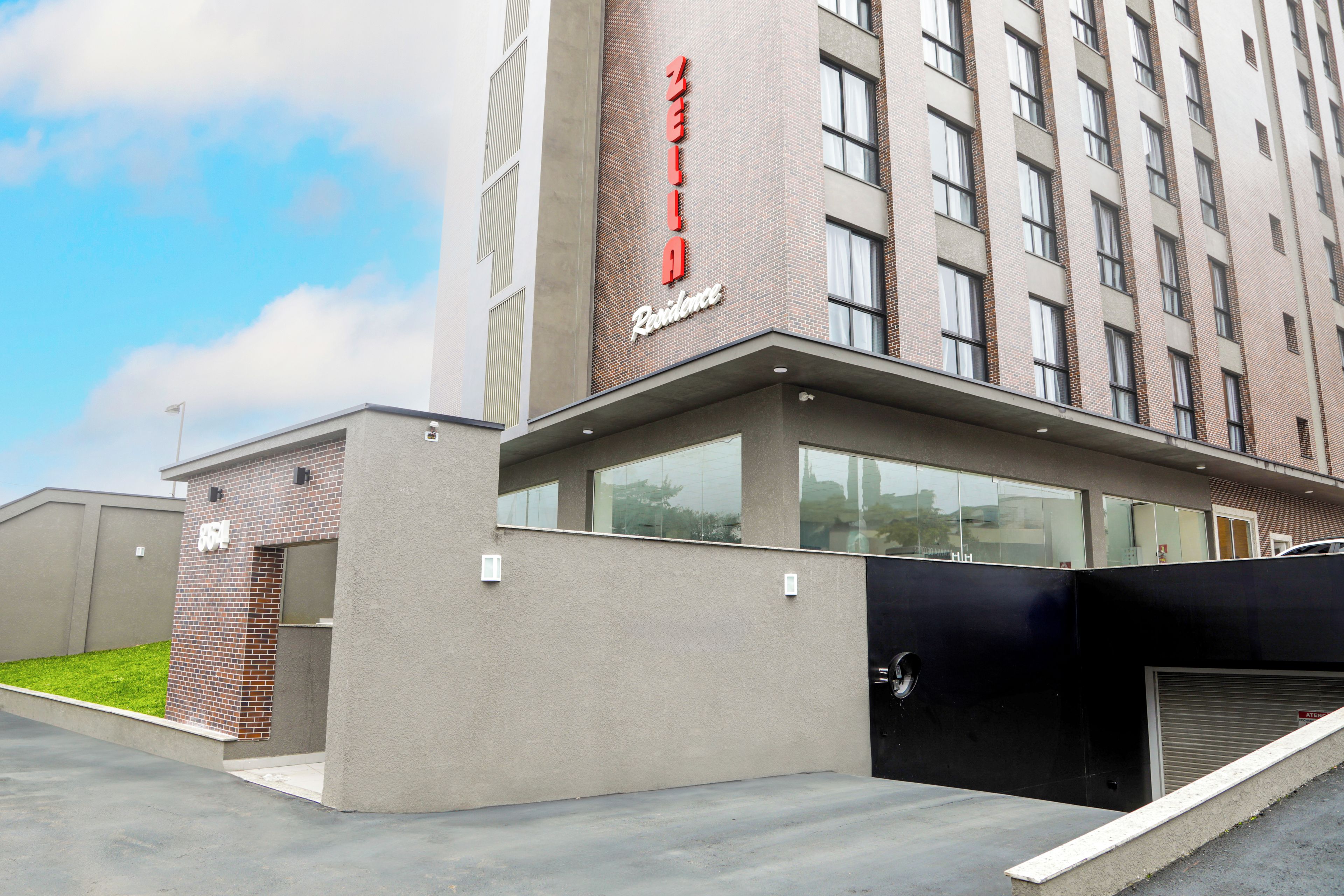 Edifício de hotel moderno com placa vermelha em destaque, entrada coberta e área de estacionamento.