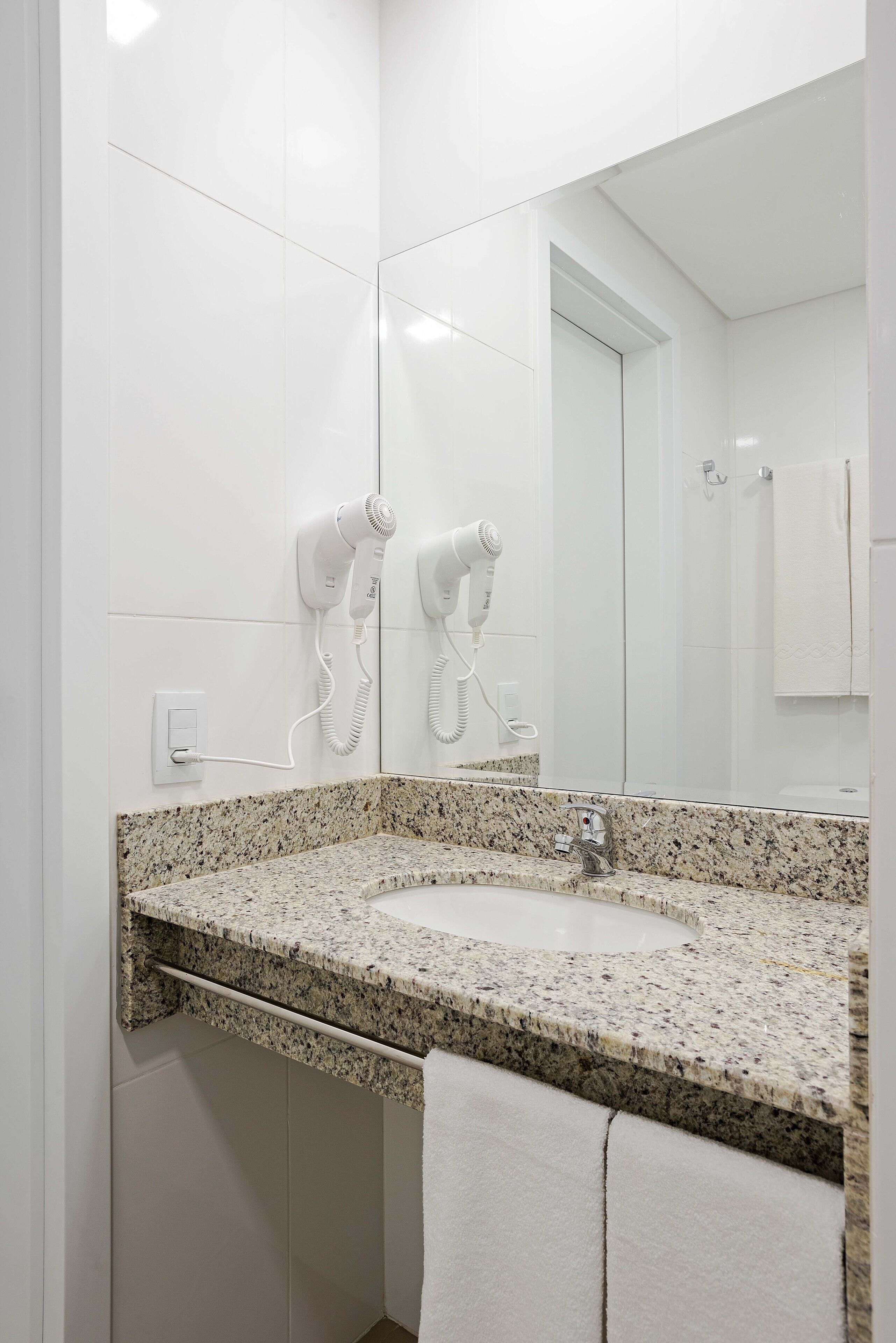 A bathroom with a granite countertop sink, white walls, and two hair dryers mounted on the wall.