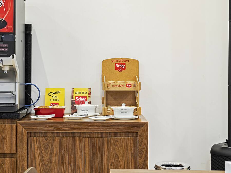 A hotel room with a wooden desk, a coffee machine, a bowl of fruit, and a sign indicating gluten-free options.