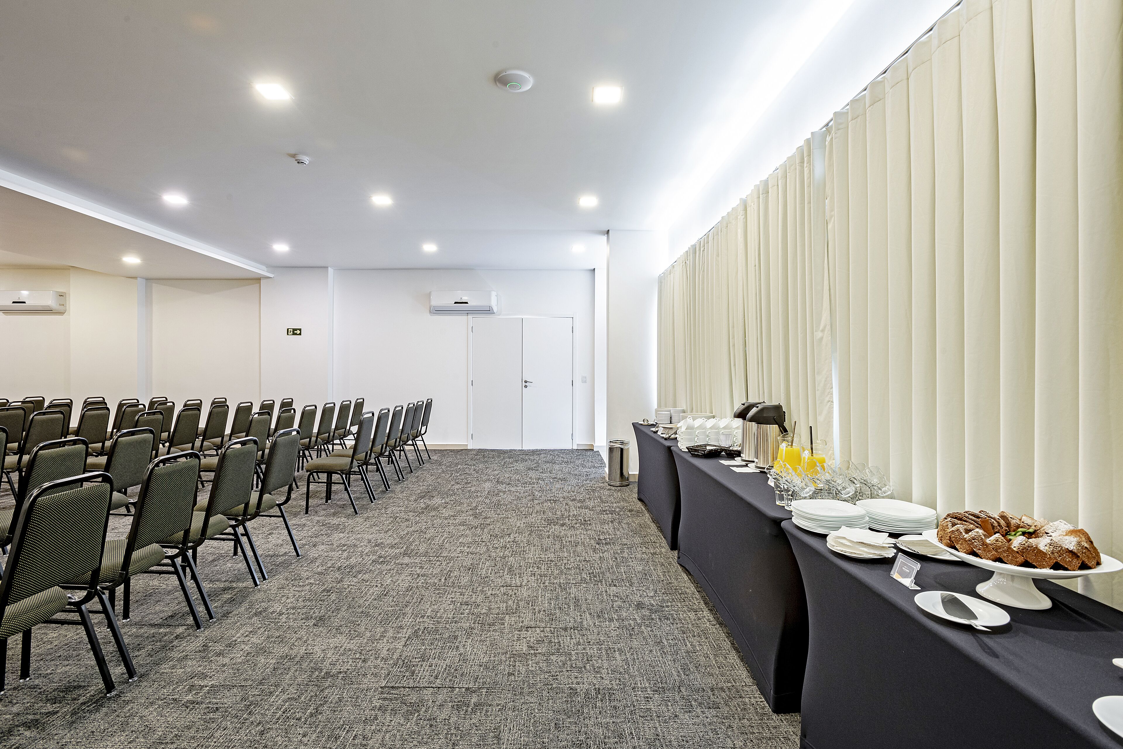 Un salón de conferencias con filas de sillas, una mesa de buffet con platos y un pastel, y un equipo de aire acondicionado en la pared.