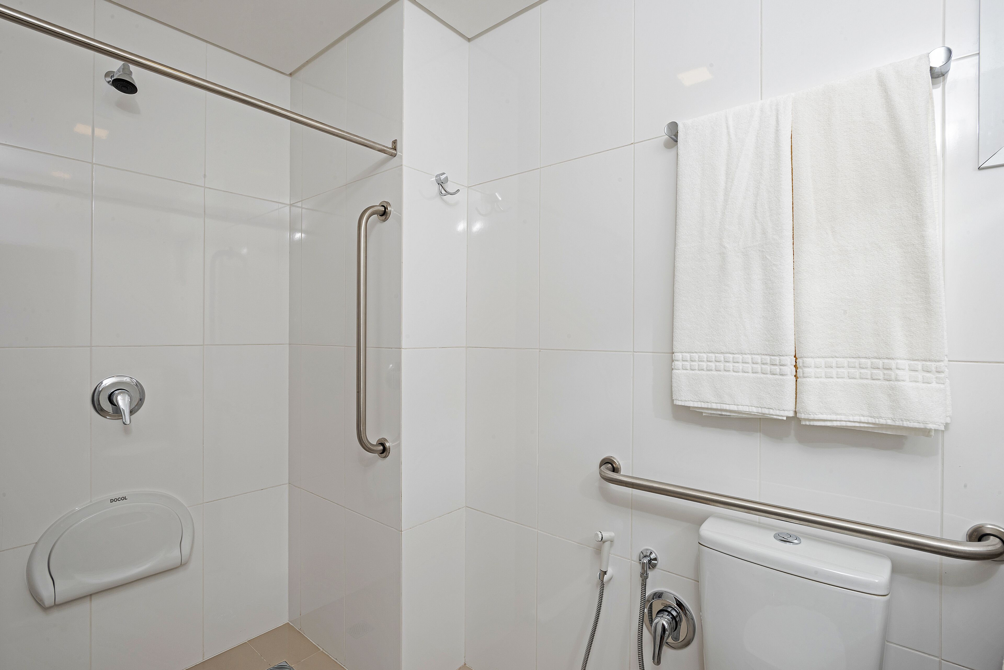 Un baño con azulejos blancos, inodoro, ducha con cabezal de ducha de mano y barras de apoyo.