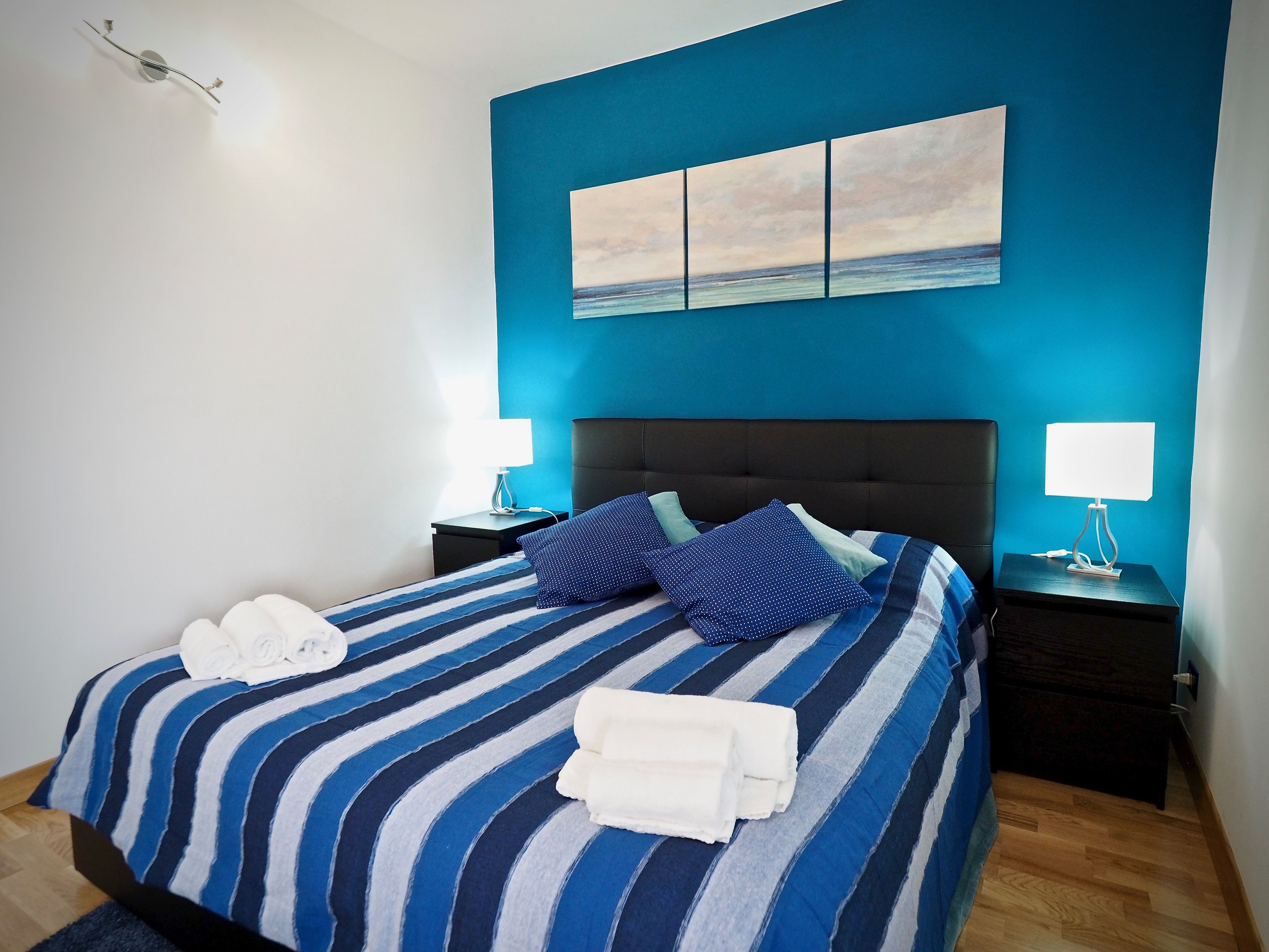 A neatly made bed with striped bedding, two pillows, and folded towels. There are two bedside tables with lamps, a wall-mounted artwork, and a blue accent wall.