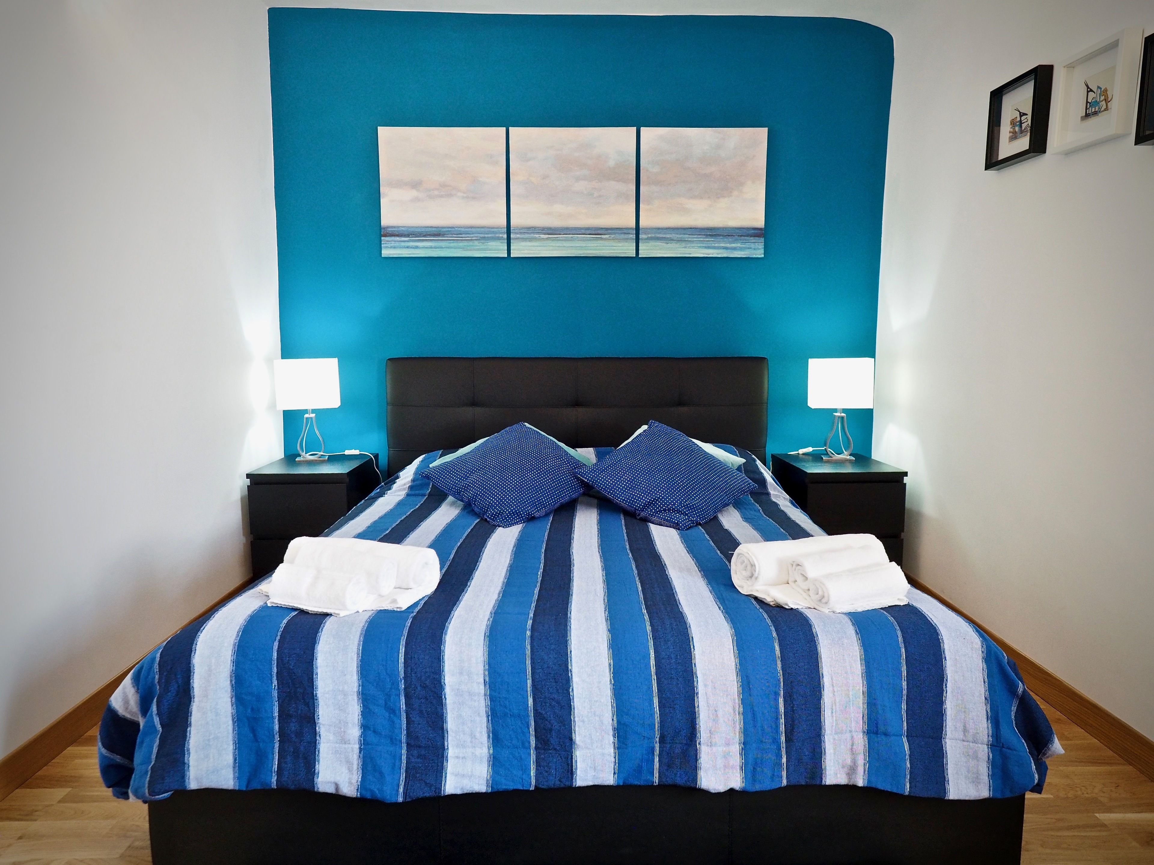A bed with a blue and white striped bedspread, two pillows, and two white towels. There are two bedside tables with lamps, a blue wall, and a painting above the bed.