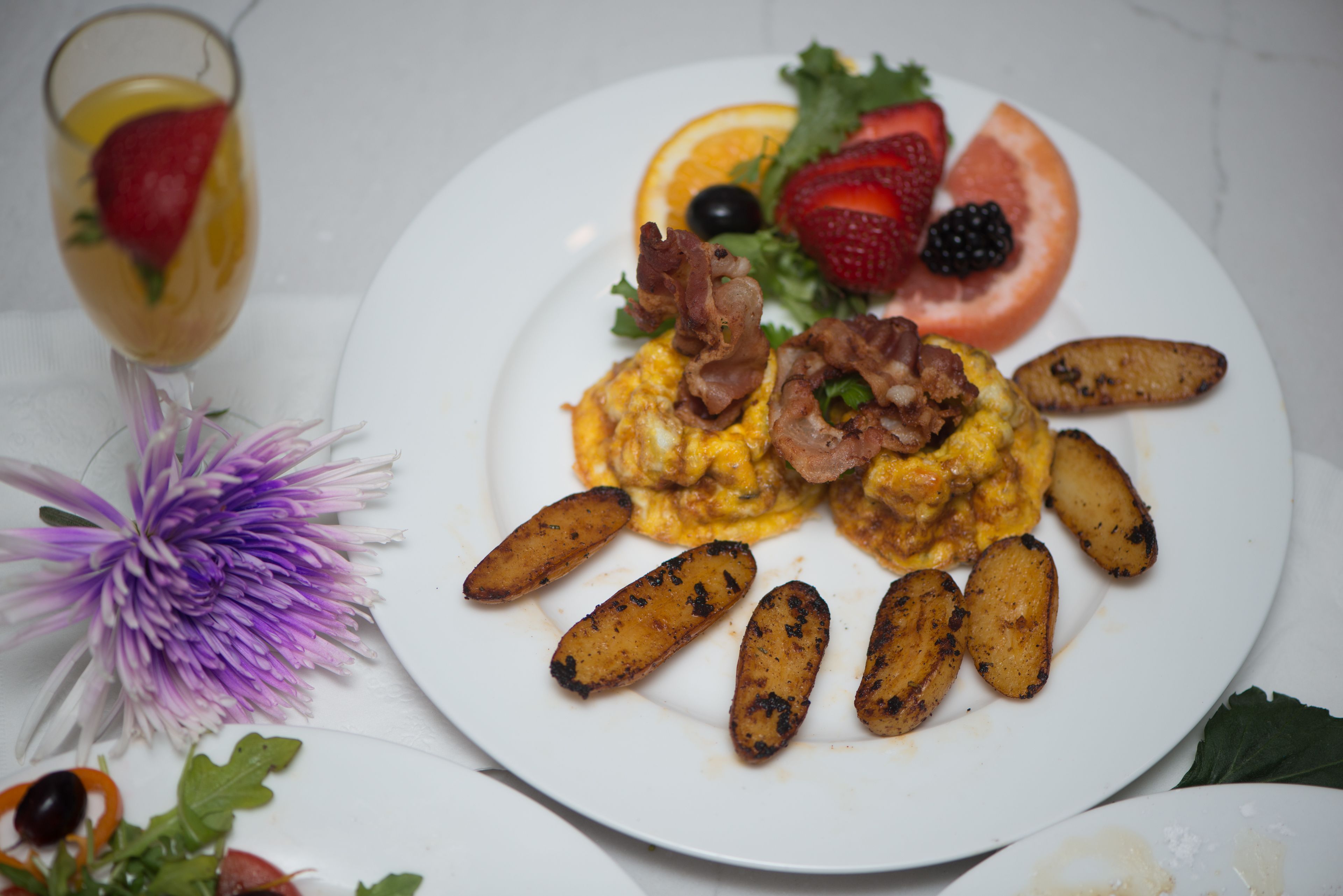 A plate with scrambled eggs topped with bacon, surrounded by roasted potatoes and garnished with strawberries, blackberries, and an orange slice.