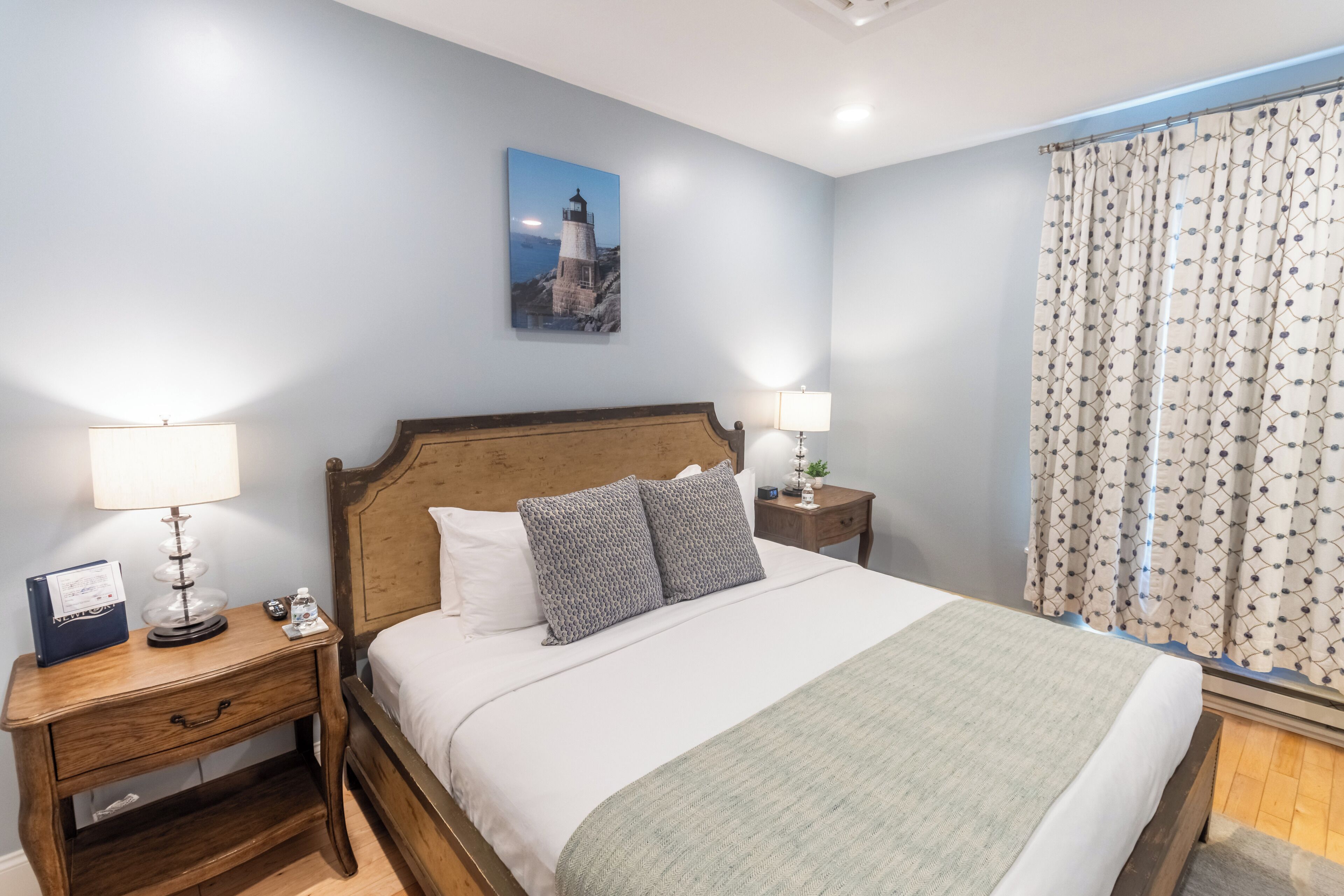 Un lit bien fait, avec une tête de lit en bois, deux tables de chevet avec des lampes, une petite table de nuit avec une lampe, un tableau encadré au mur et une fenêtre avec des rideaux.