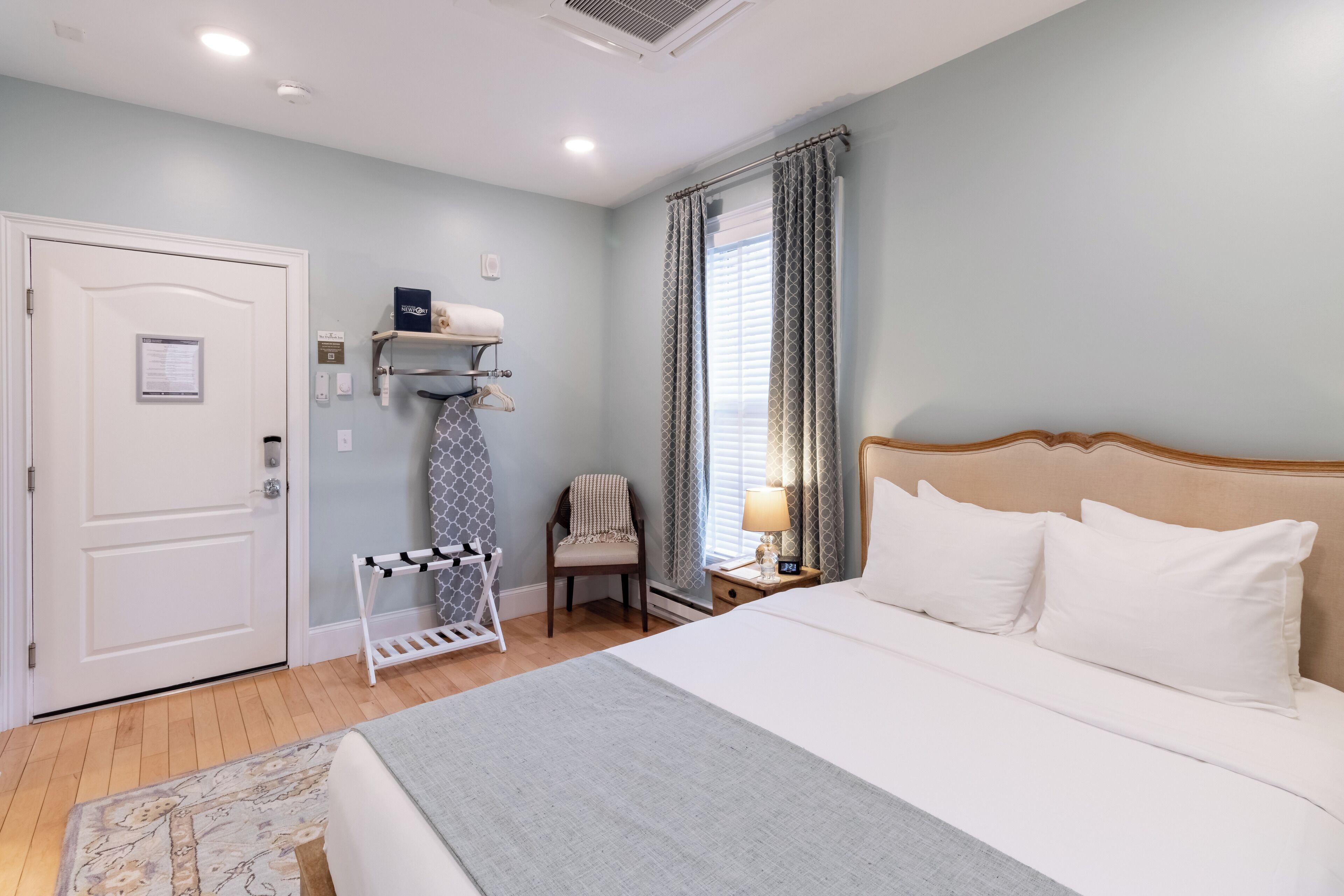 A hotel room with a bed, a chair, a nightstand, a door, and a window with curtains.