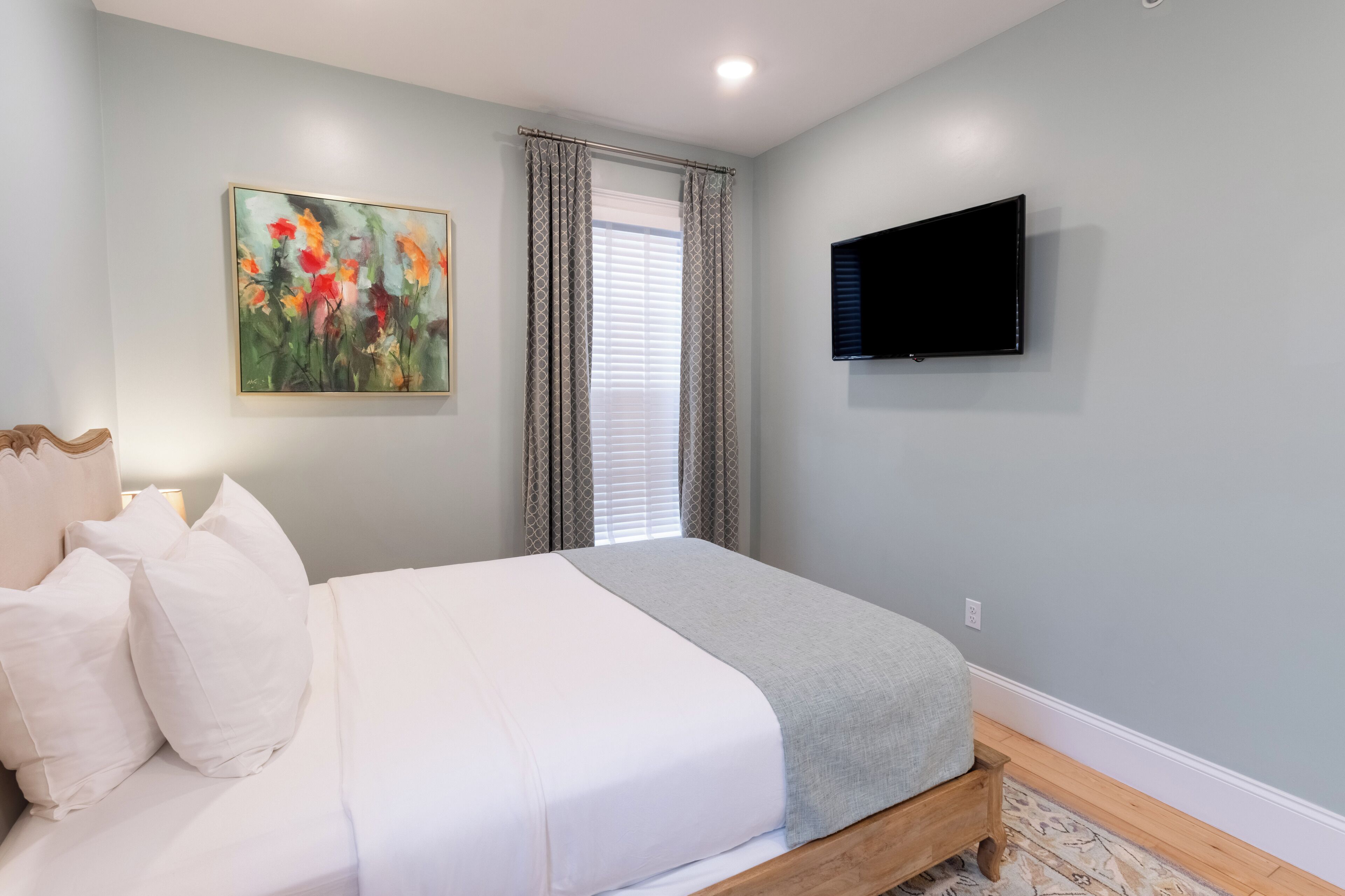 A bedroom with a bed, a painting, a TV, and a window with curtains.
