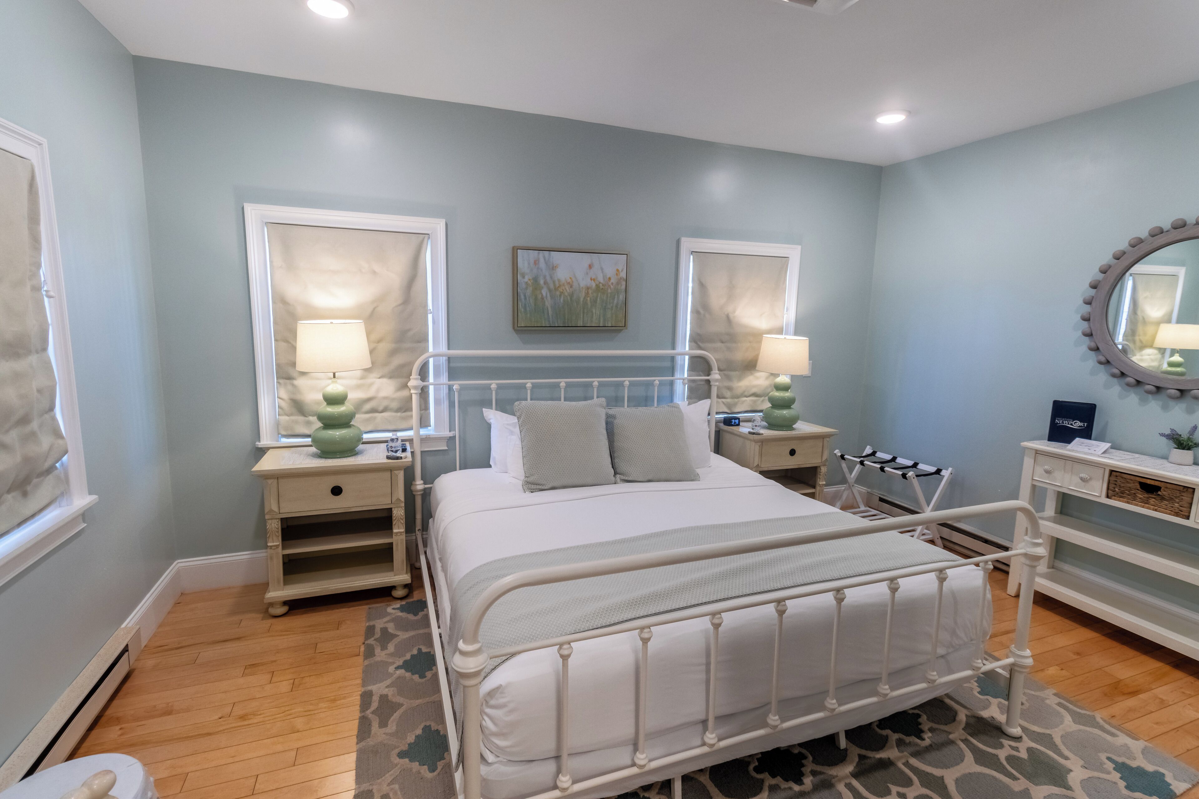 Quarto com uma cama de metal branca, duas mesas de cabeceira com luminárias, uma mesinha de cabeceira com abajur verde, um espelho e um quadro na parede.