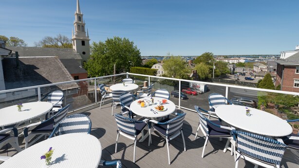 An outdoor patio with round tables and blue and white wicker chairs, overlooking a town with a church steeple. - The Outlook Inn (Newport)