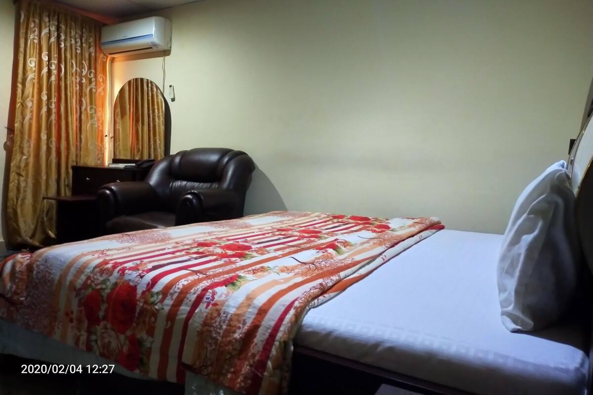 A hotel room with a bed, a chair, an air conditioning unit, and curtains.