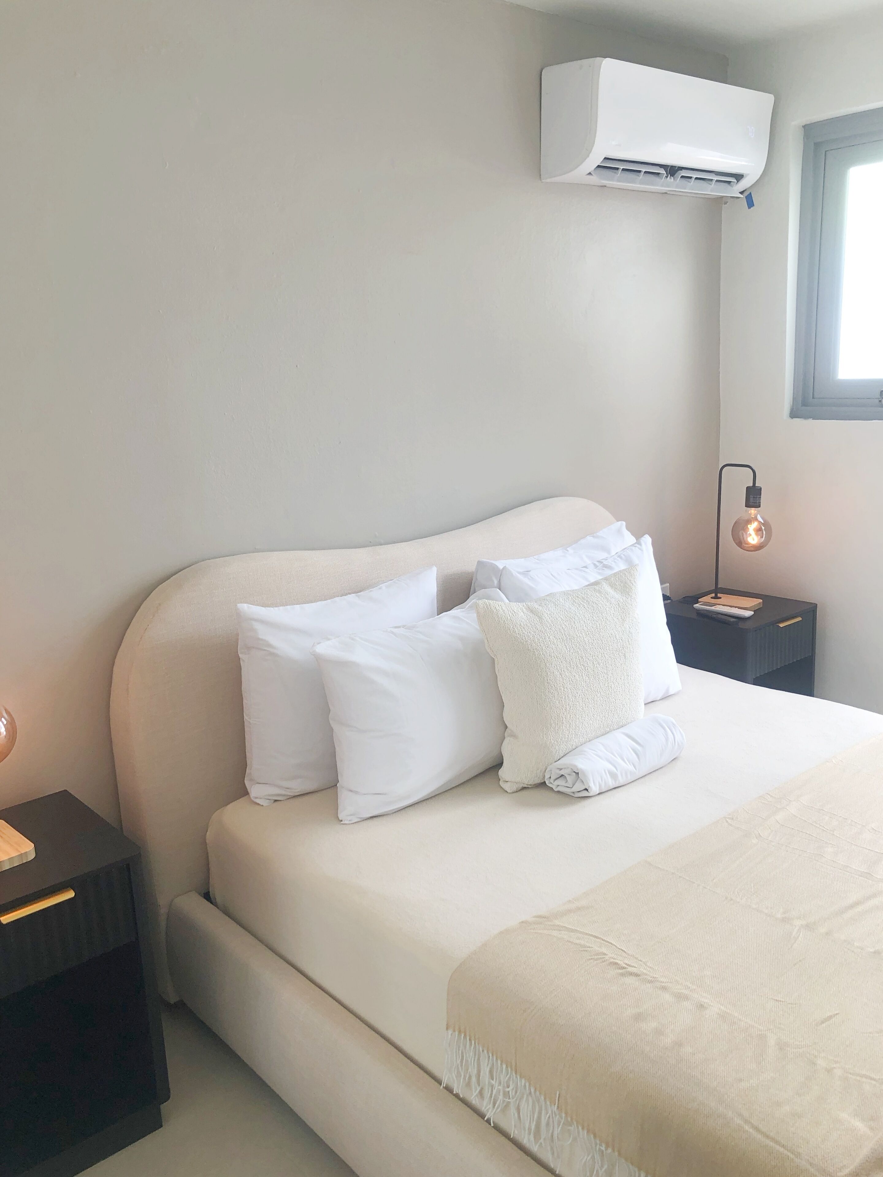A bedroom with a bed, white pillows, a bedside table, a nightstand, a lamp, and an air conditioning unit.